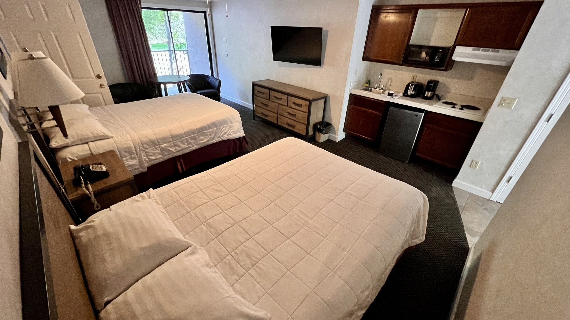 Habitación de hotel con dos camas, un escritorio con lámpara, una mesita, televisión, kitchenette y ventana con cortinas.