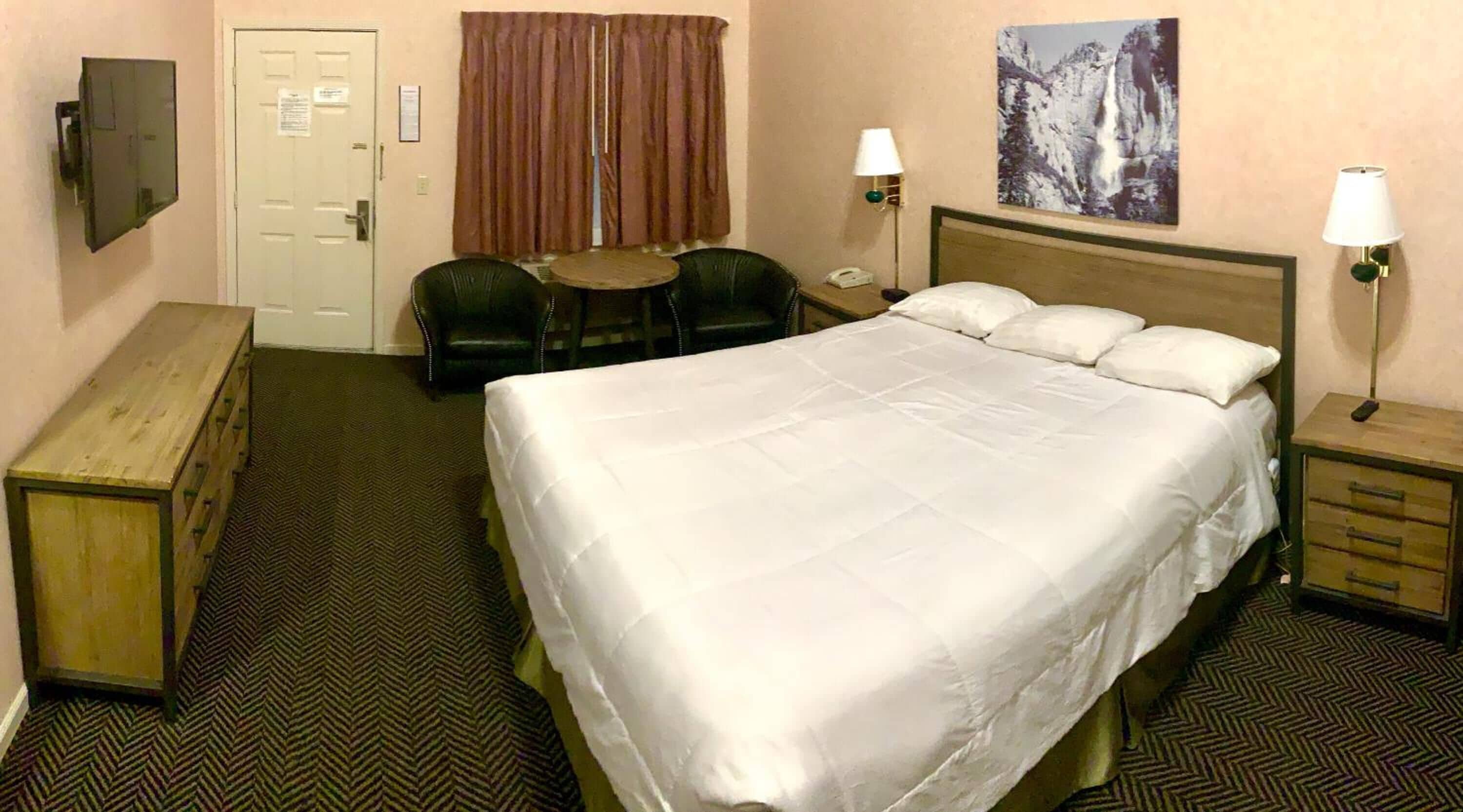 Habitación de hotel con una cama grande, un escritorio, una silla, una mesita, un televisor y un cuadro en la pared.