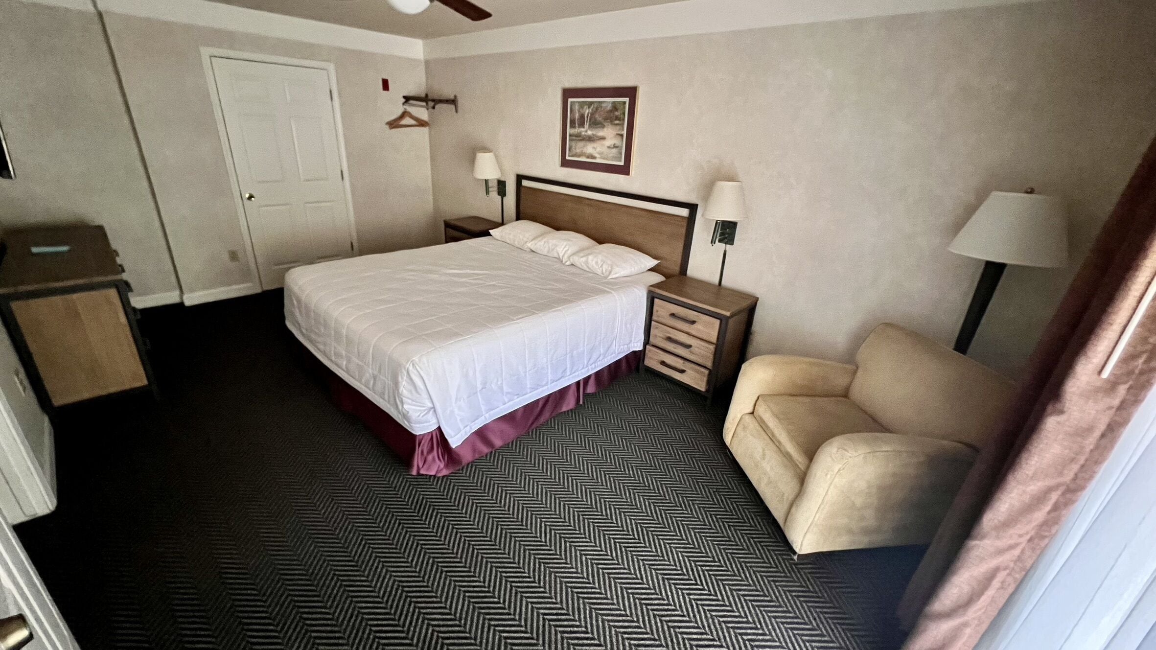 Une chambre d’hôtel avec un lit, des tables de chevet, une chaise, une porte et un tableau encadré au mur.