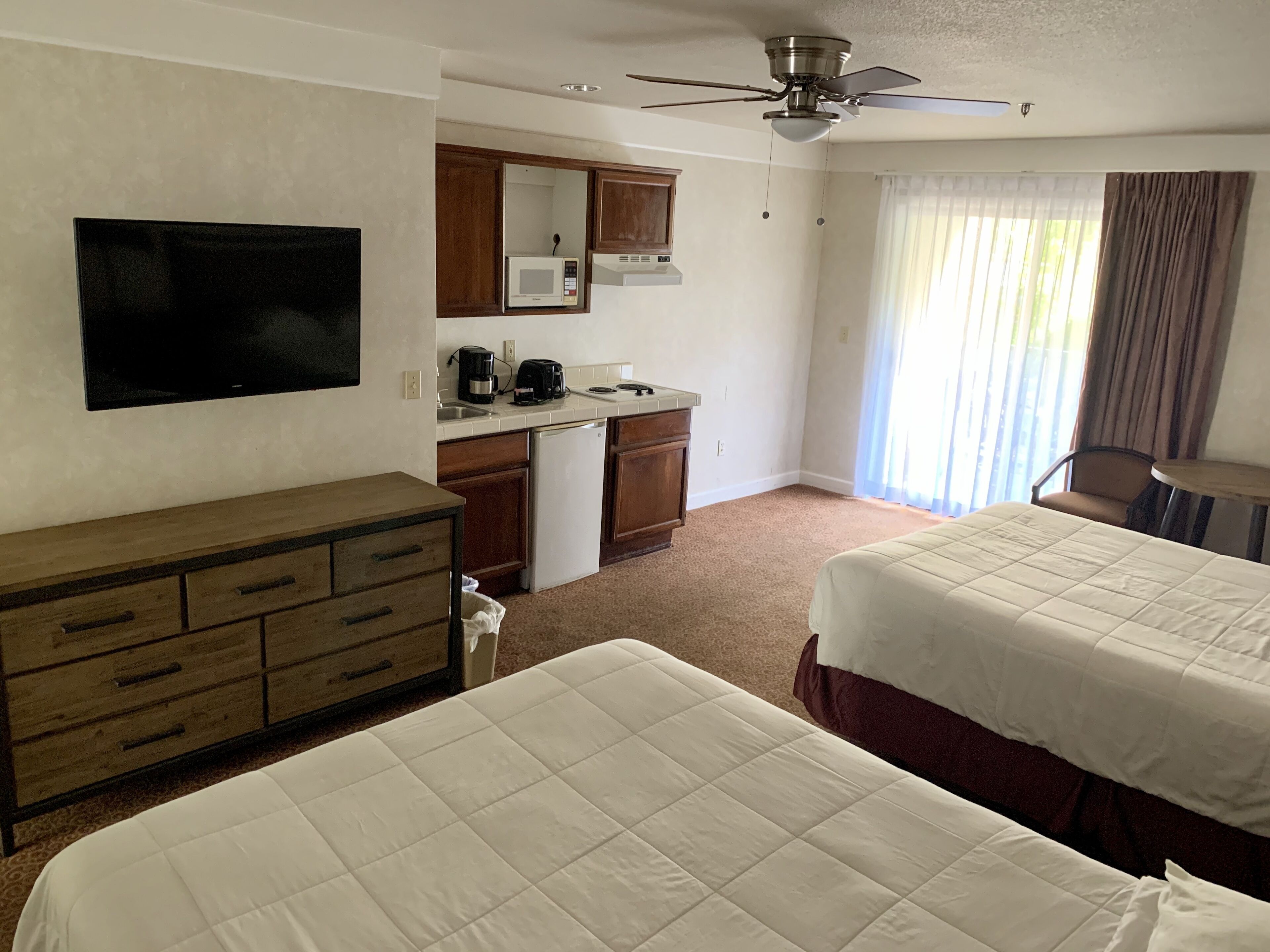 Une chambre d’hôtel avec deux lits, une kitchenette, un ventilateur de plafond et une grande fenêtre avec des rideaux.