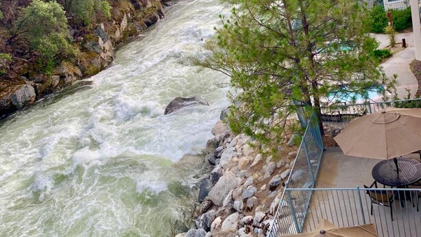 A river with a rocky riverbank, a tree, and outdoor furniture with an umbrella. - Yosemite View Lodge (El Portal)