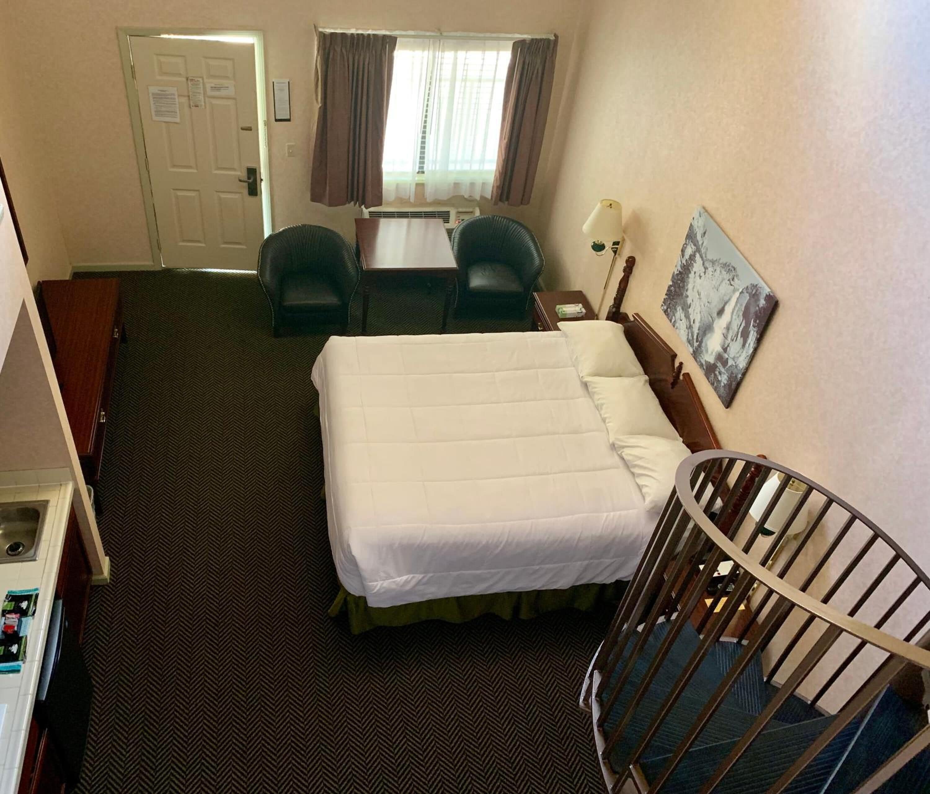 A hotel room with a bed, a desk, two chairs, a small table, a nightstand, a lamp, a painting, and a staircase.