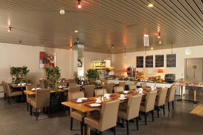 A spacious dining area with multiple tables and chairs, a kitchen in the background, and a ceiling with linear lighting fixtures.