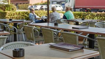 Un café en plein air avec des chaises et des tables en osier, un cendrier noir et un livre posé sur une table.