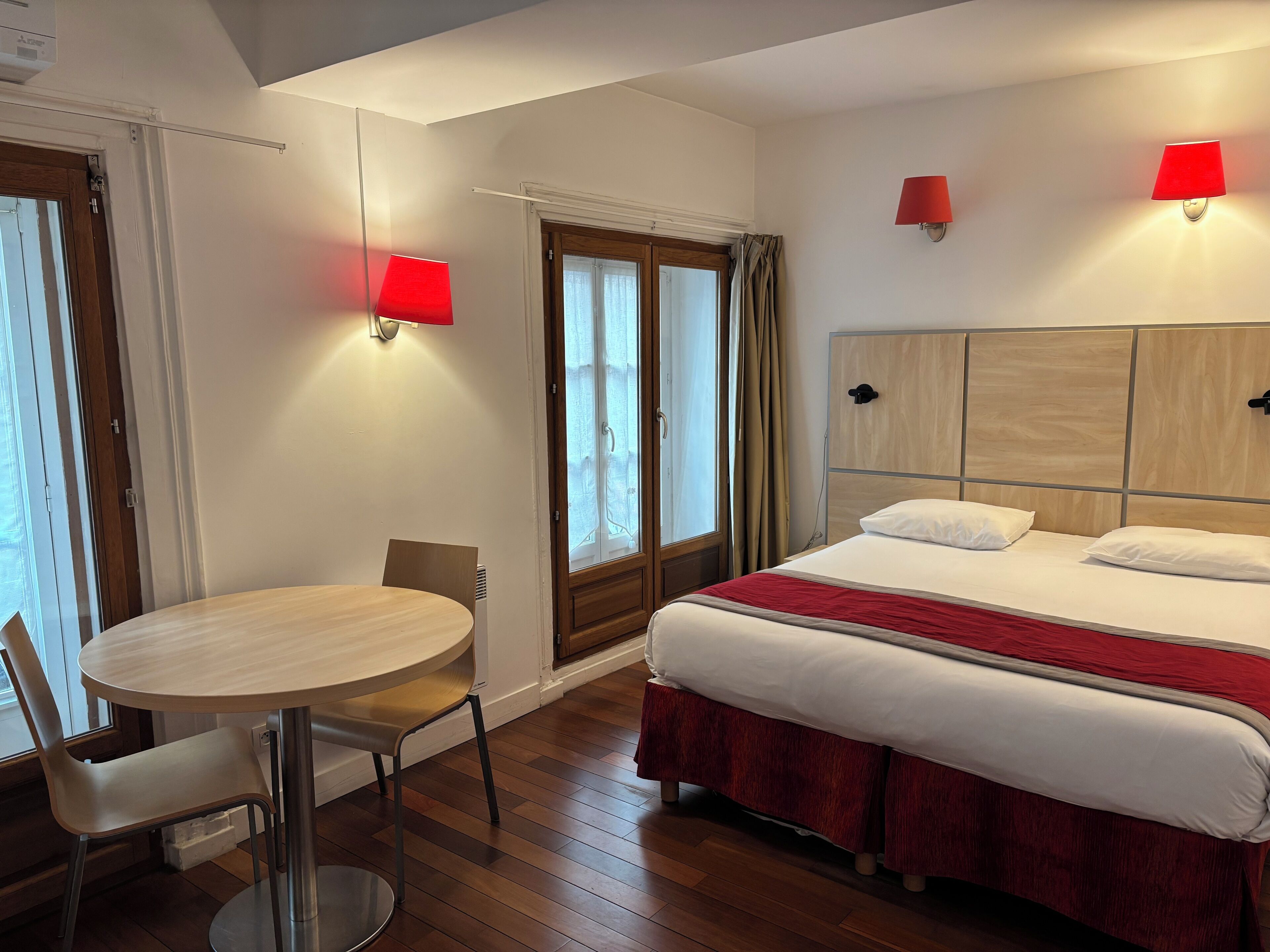 A hotel room with a bed, a round table with a chair, a window with curtains, and two wall-mounted lamps.