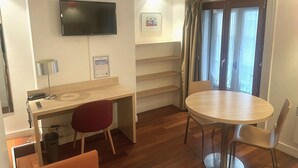 Una habitación de hotel compacta con un escritorio de madera, un televisor montado en la pared, una pequeña mesa con dos sillas y una ventana con cortinas.