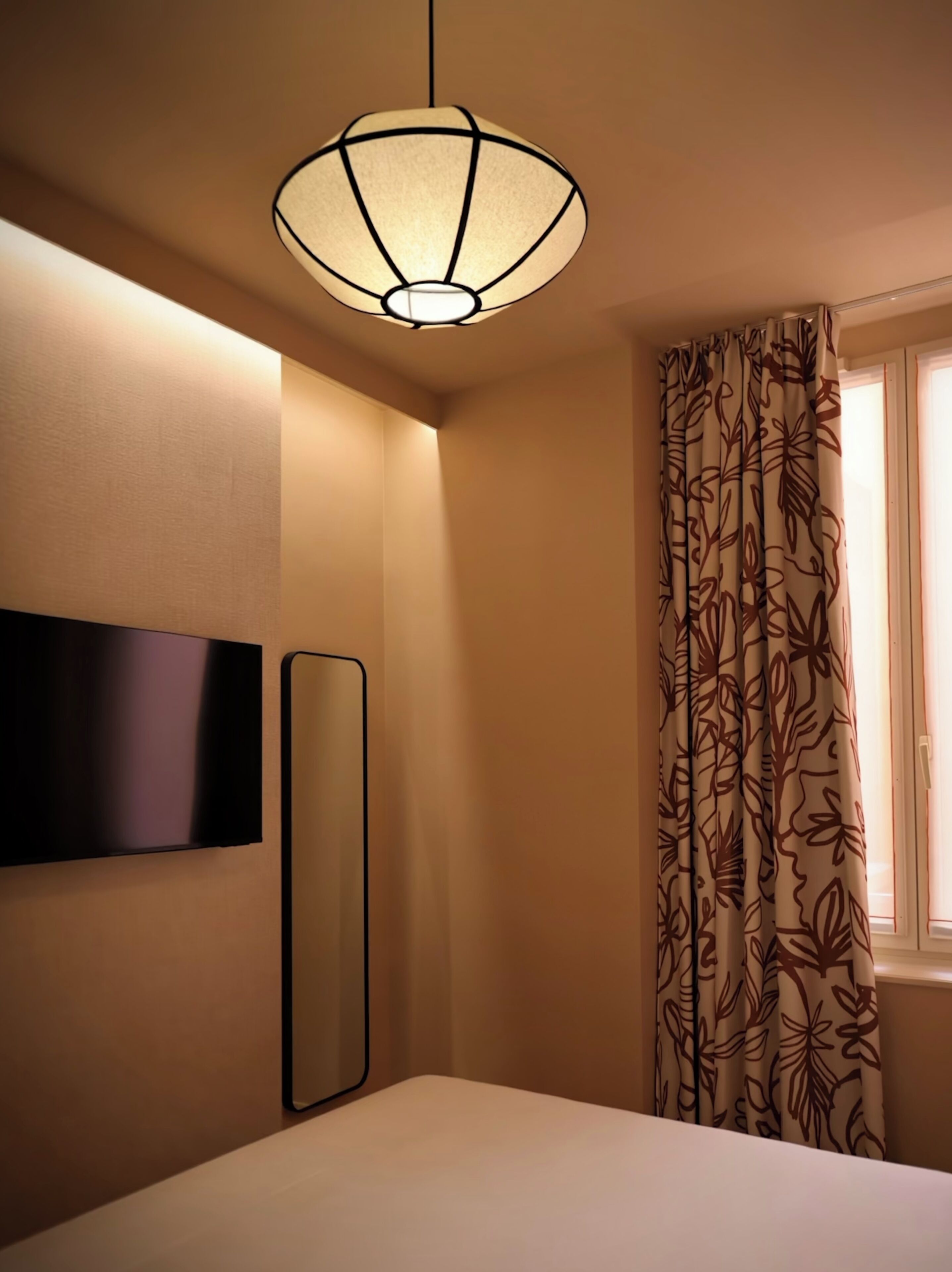 Ein Hotelzimmer mit Bett, einem an der Wand montierten Fernseher, einer modernen Pendelleuchte und einem mit Vorhängen versehenen Fenster.