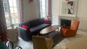 Una sala de estar con un sofá, una mesa de centro y dos sillones. Hay una chimenea y una ventana con cortinas.