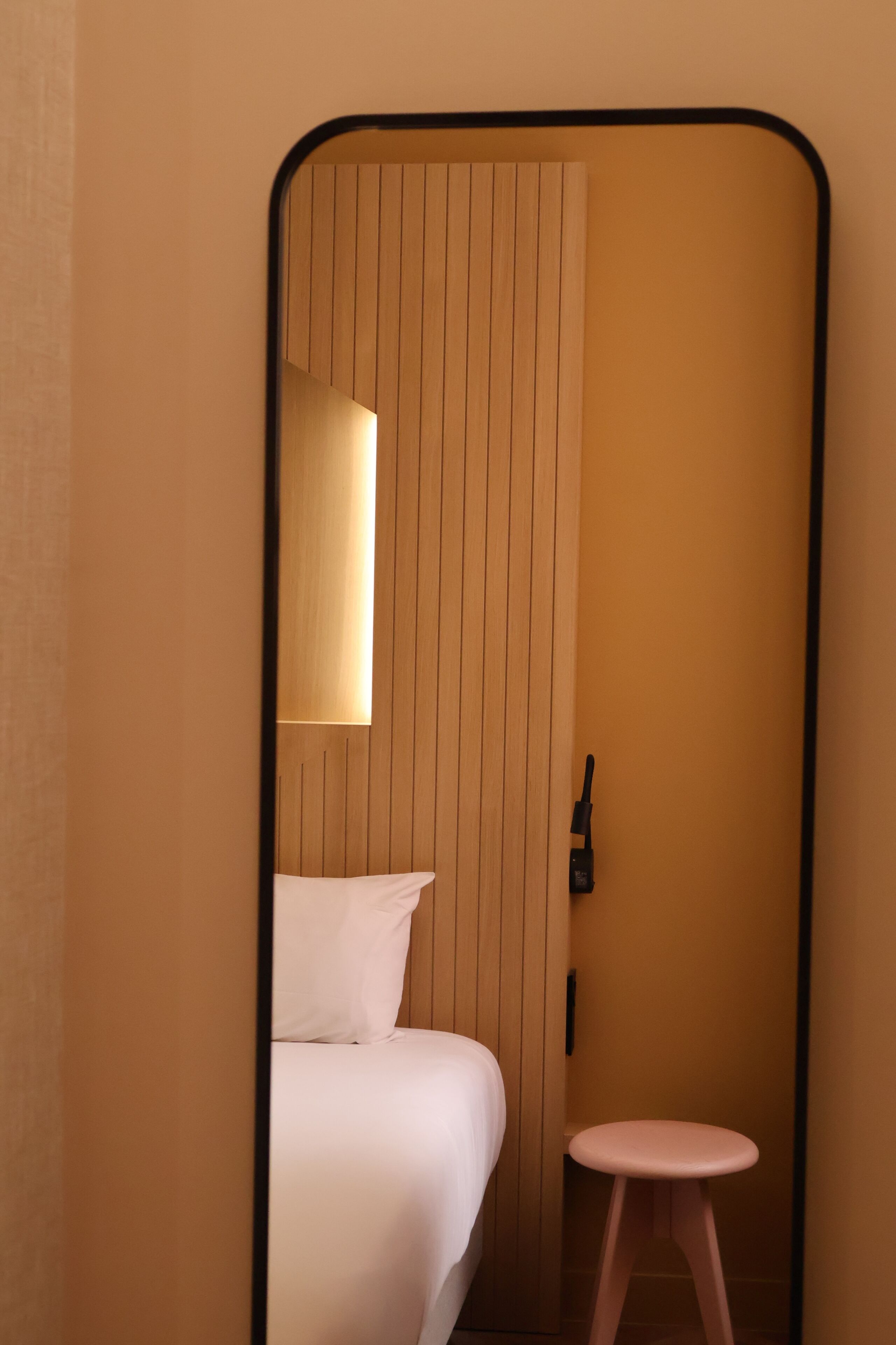 Une chambre d’hôtel avec un lit, un petit tabouret, une table de chevet et une lampe fixée au mur.