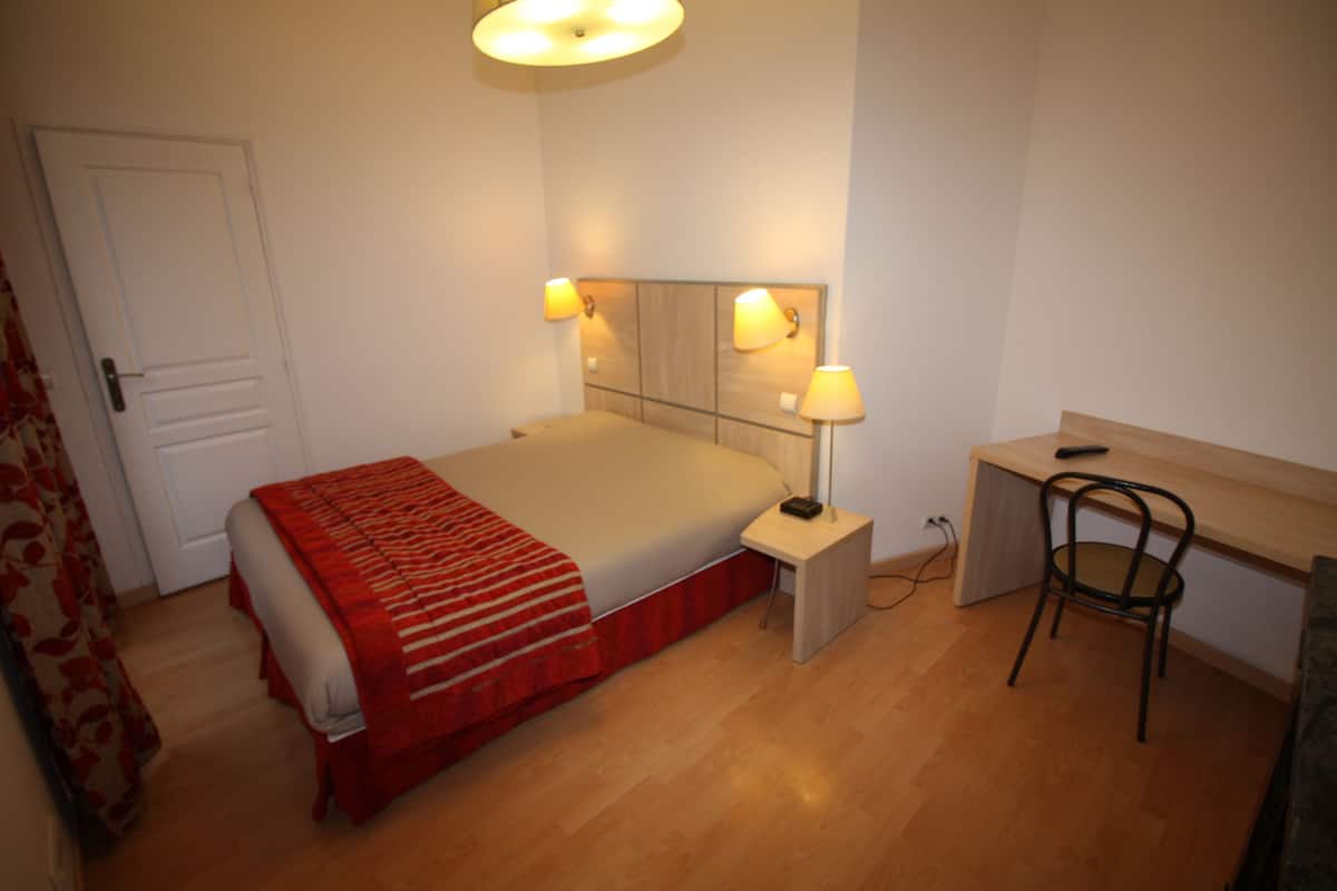 A bedroom with a bed, bedside table, lamp, chair, and desk.