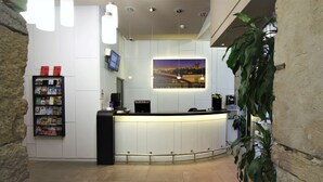 Zona de recepción con mostrador curvo, pantalla digital, máquina de café y estantería con revistas.