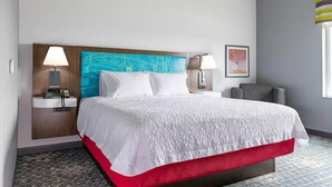 A hotel room with a large bed, two bedside lamps, a headboard with a blue artwork, a small table with a lamp, and a window with a view. - Hampton Inn by Hilton Chelsea (Chelsea)