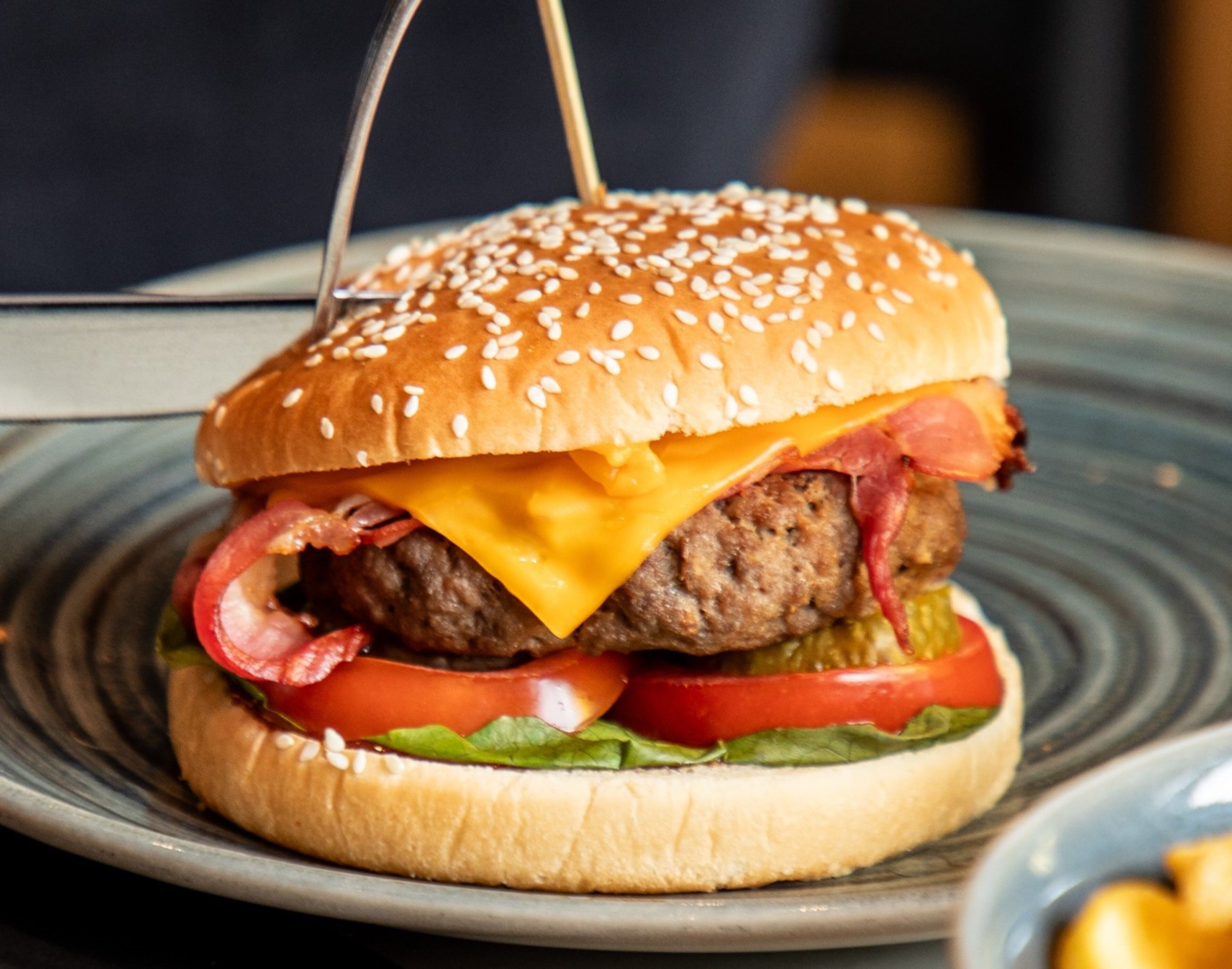 A burger with sesame seed bun, cheese, bacon, lettuce, tomato, and a pickled vegetable, served on a plate.