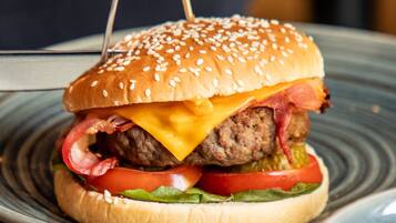 A burger with sesame seed bun, cheese, bacon, lettuce, tomato, and a pickled vegetable, served on a plate.