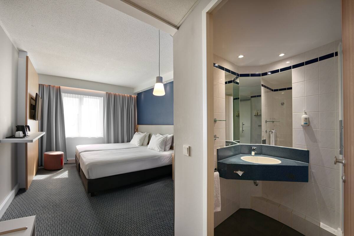 A hotel room with a bed, a desk with a chair, a bathroom with a sink and mirror, and a shower area.