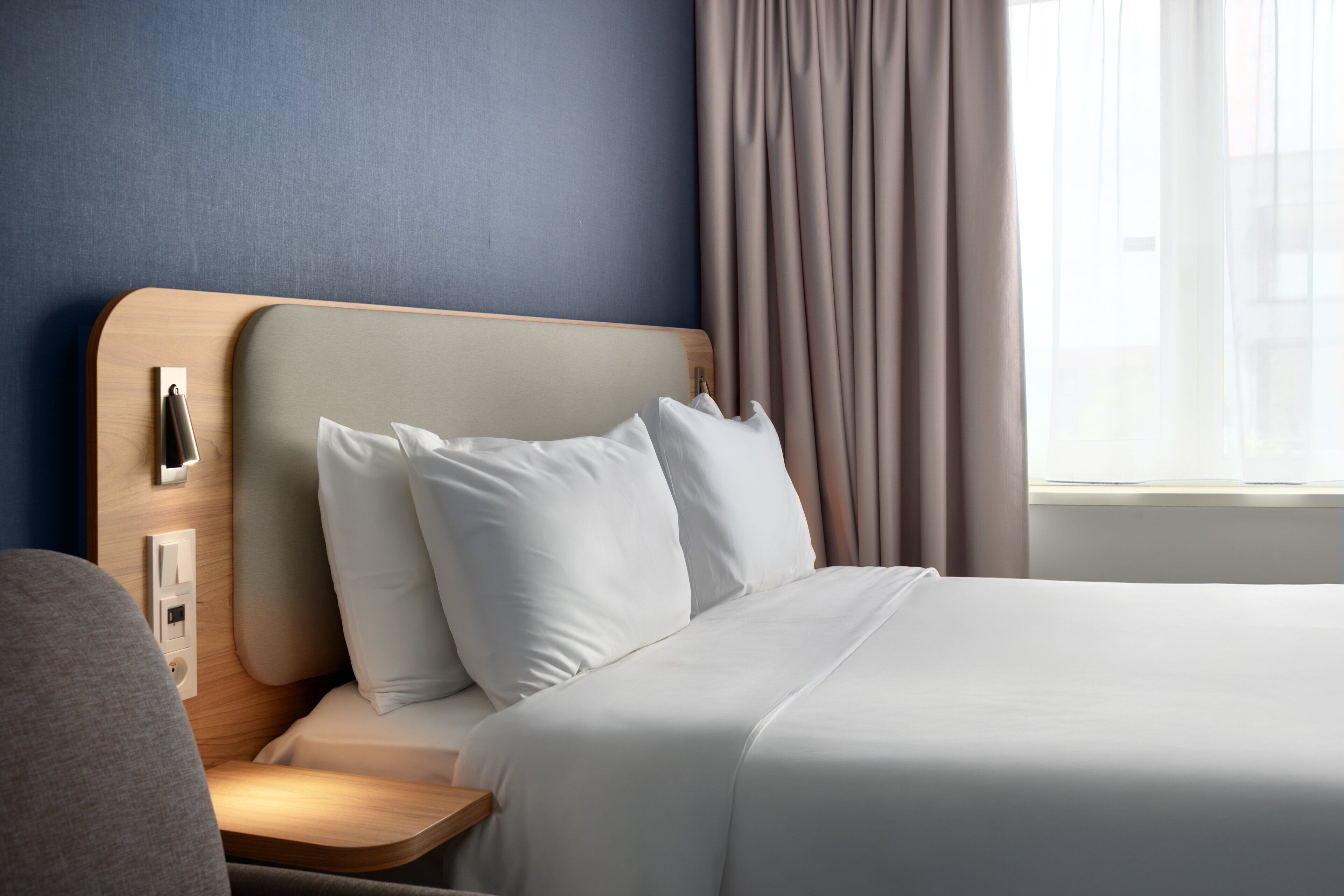 A hotel room with a neatly made bed, a bedside table, a nightstand, a lamp, and a window with curtains.