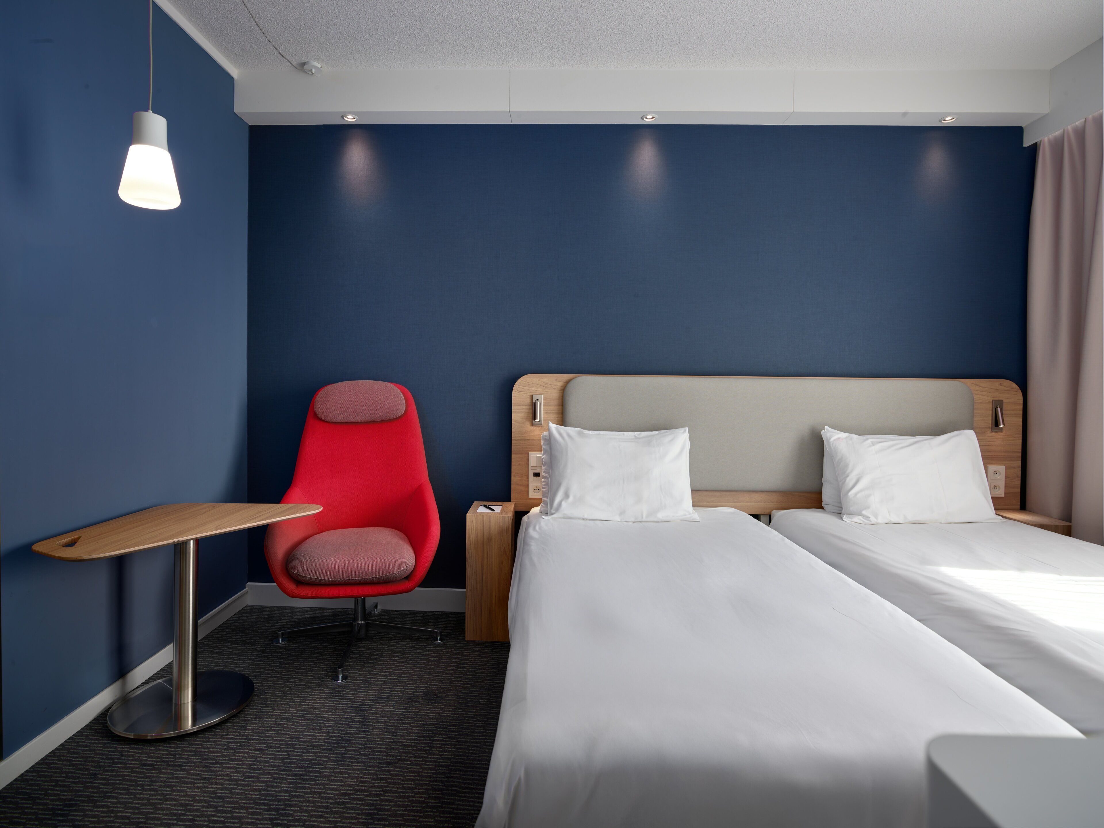 A hotel room with two beds, a red chair, a small table, and a ceiling light.