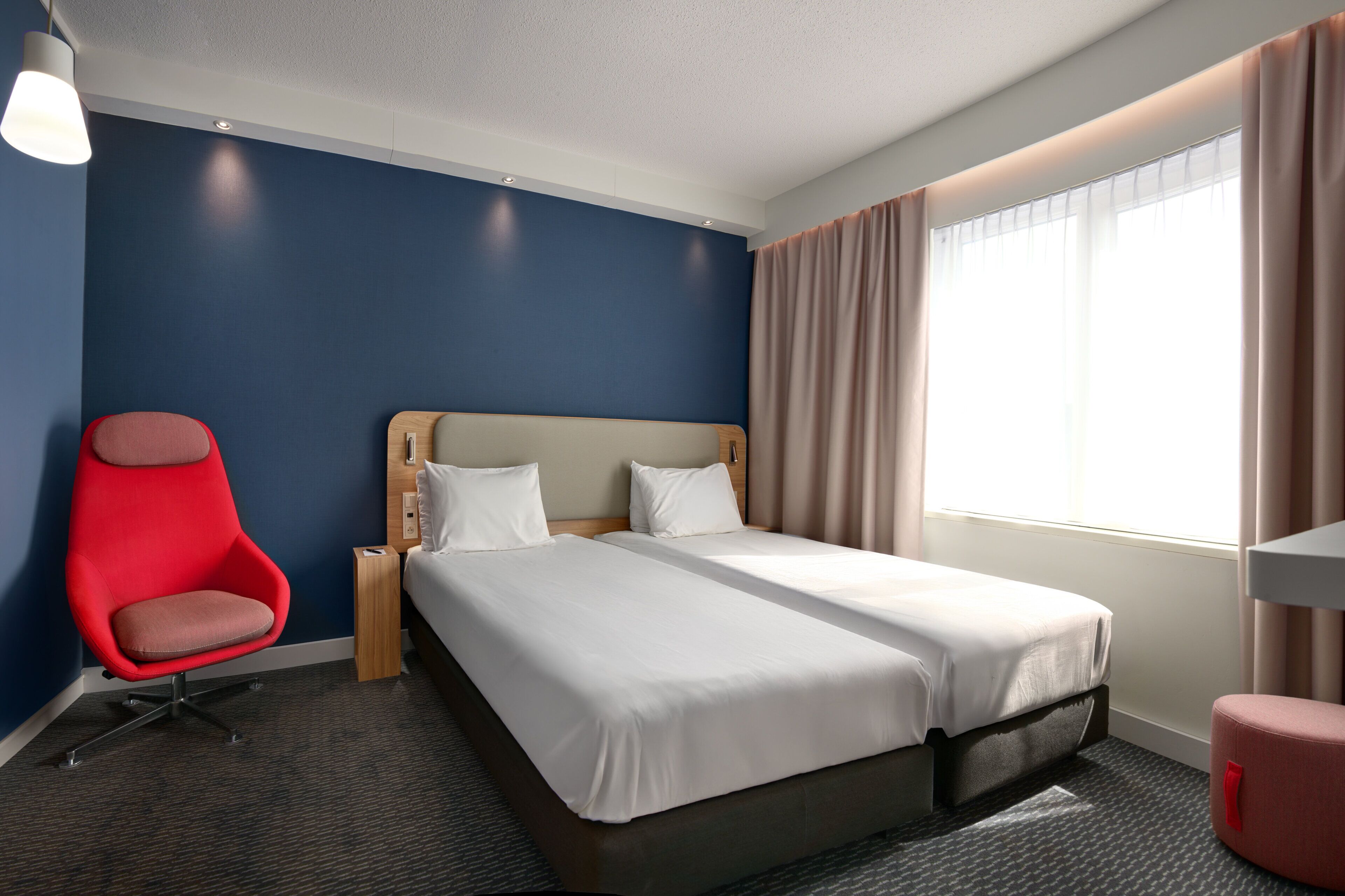 Een hotelkamer met een groot bed, een rode stoel, een klein tafeltje en een raam met gordijnen.