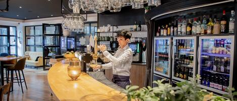 A bar area with a bartender preparing drinks, a wooden counter, and a refrigerator stocked with bottles.