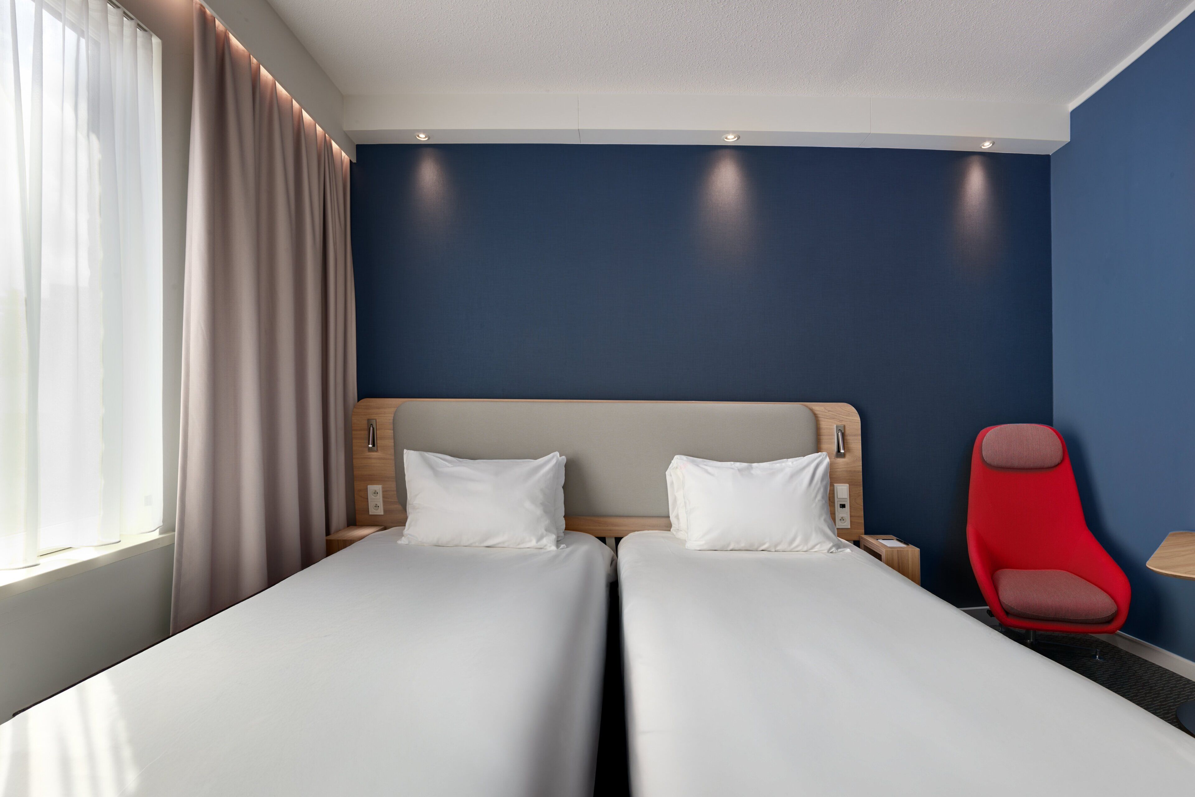 A hotel room with two beds, a red chair, and a nightstand.