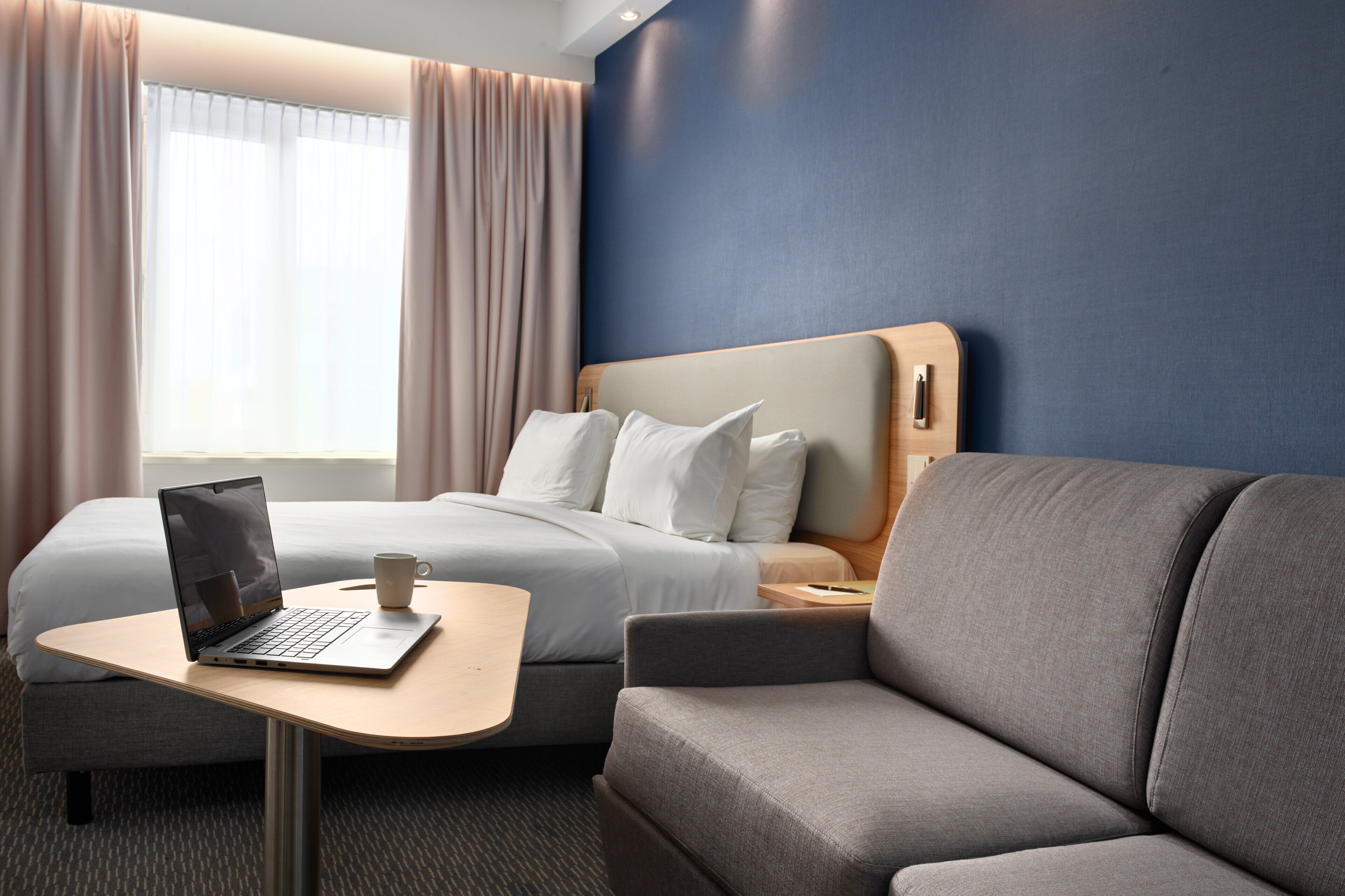 Une chambre d’hôtel avec un lit, un canapé, une table avec un ordinateur portable et une tasse, et une fenêtre avec des rideaux.