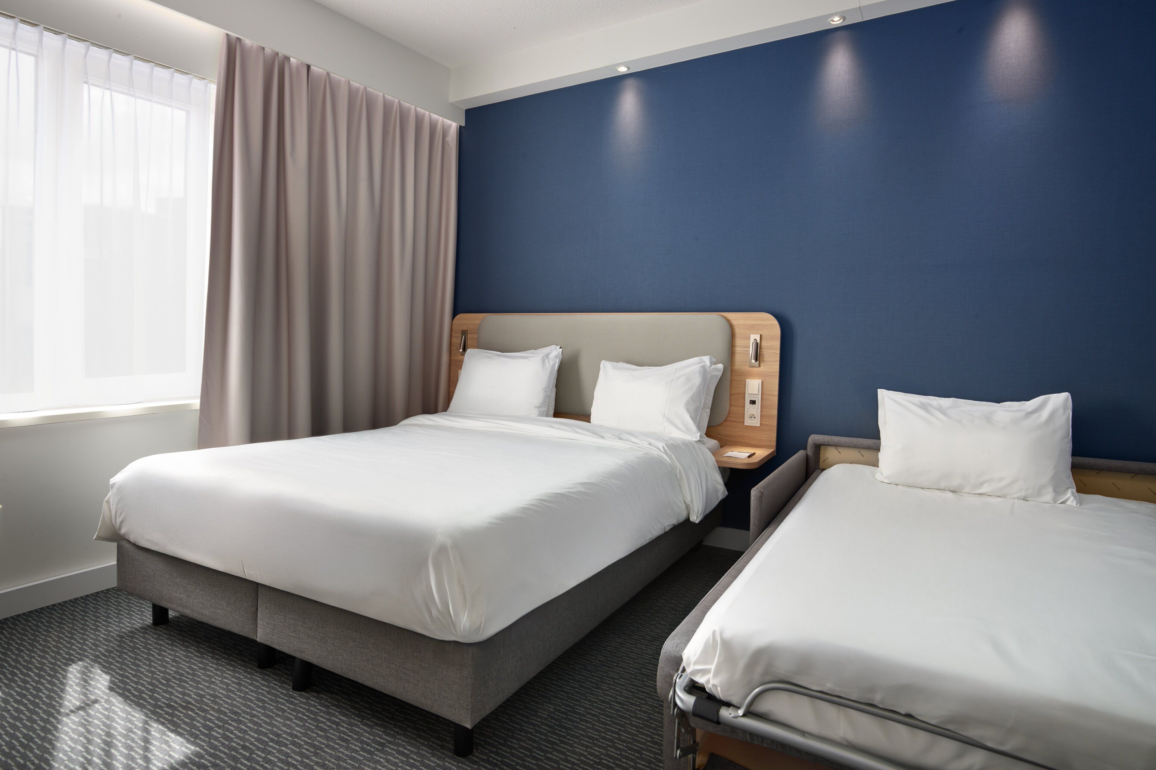 Habitación de hotel con dos camas, un ventanal con cortinas, una pared de acento azul y un cabecero de madera.