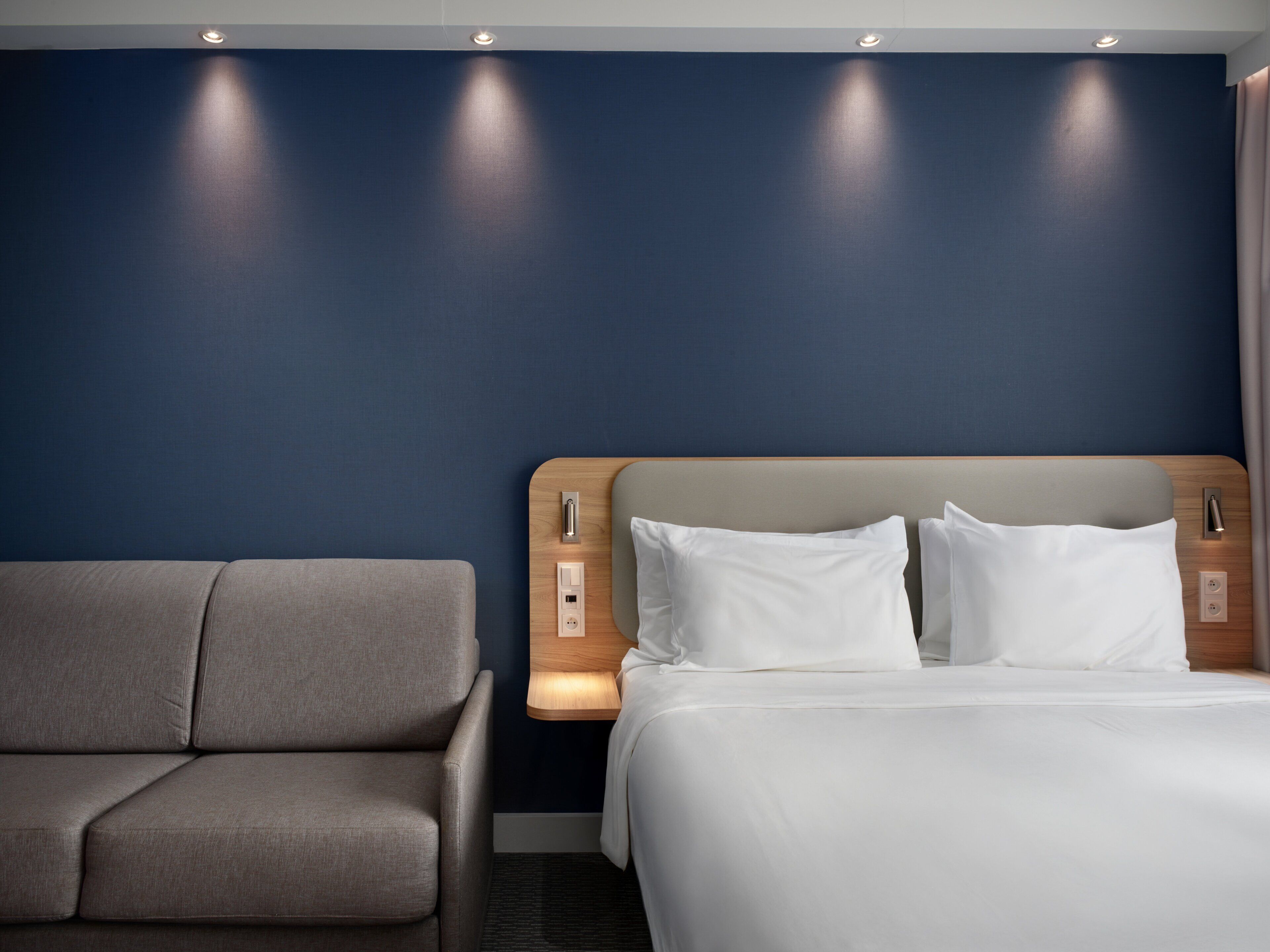 A hotel room with a bed, a sofa, and a wall with recessed lighting.