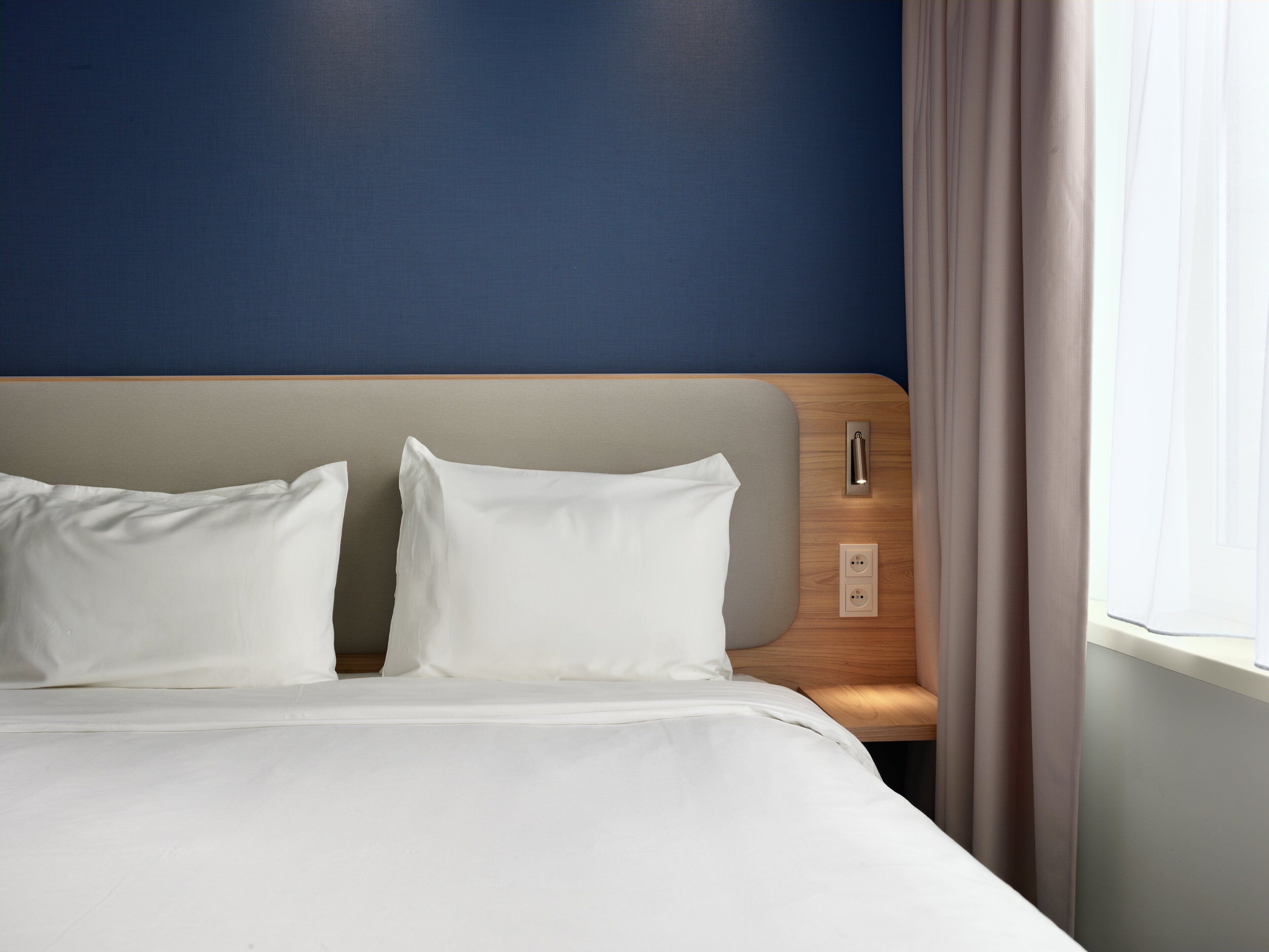 Een hotelkamer met een bed, hoofdeinde, nachtkastje en een raam met gordijnen.