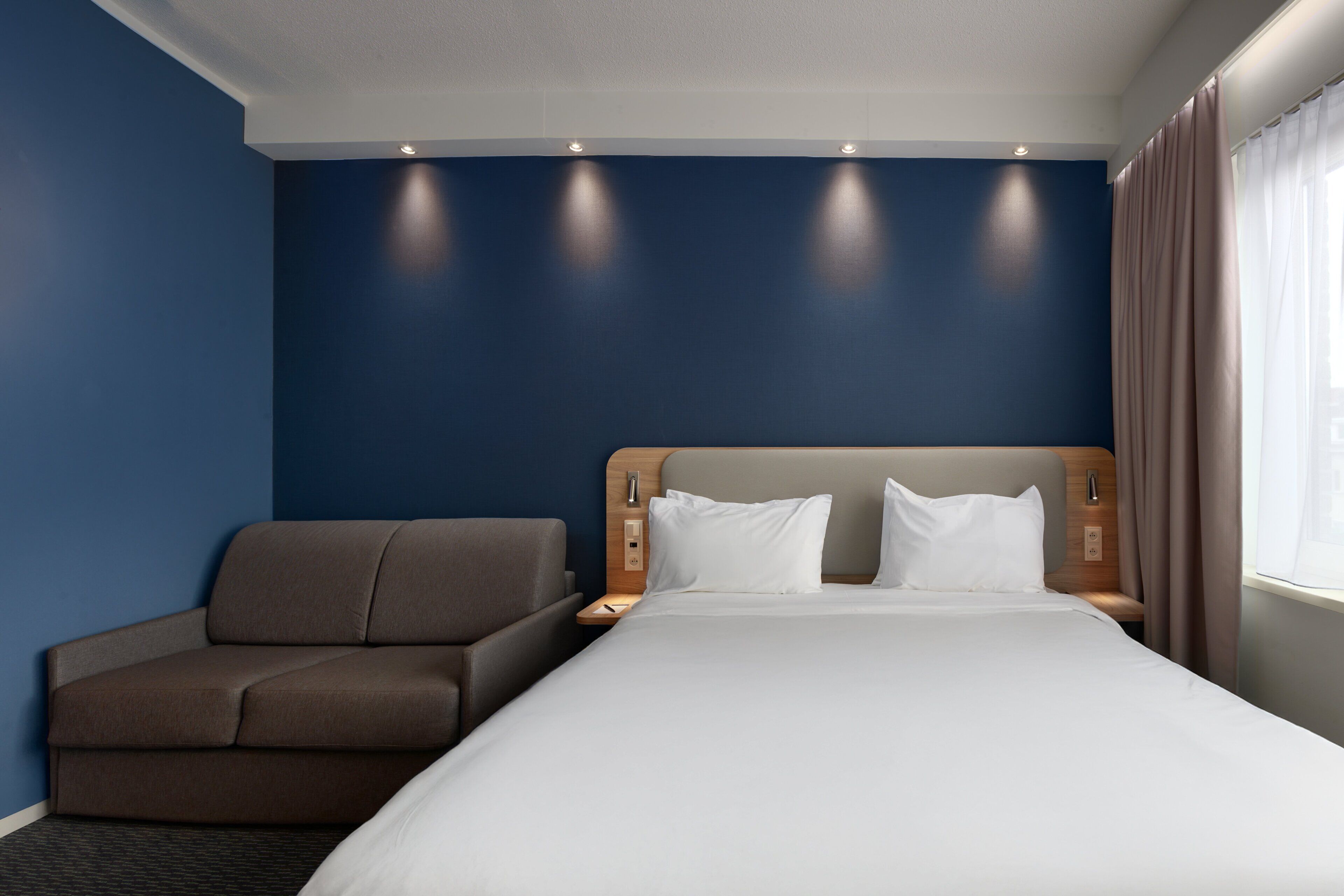 A hotel room with a bed, a sofa, a nightstand, and a window with curtains.