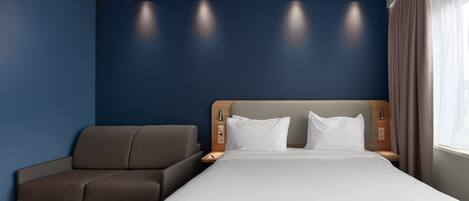 Una habitación de hotel con una cama, un sofá, una mesita de noche y un ventanal con cortinas.