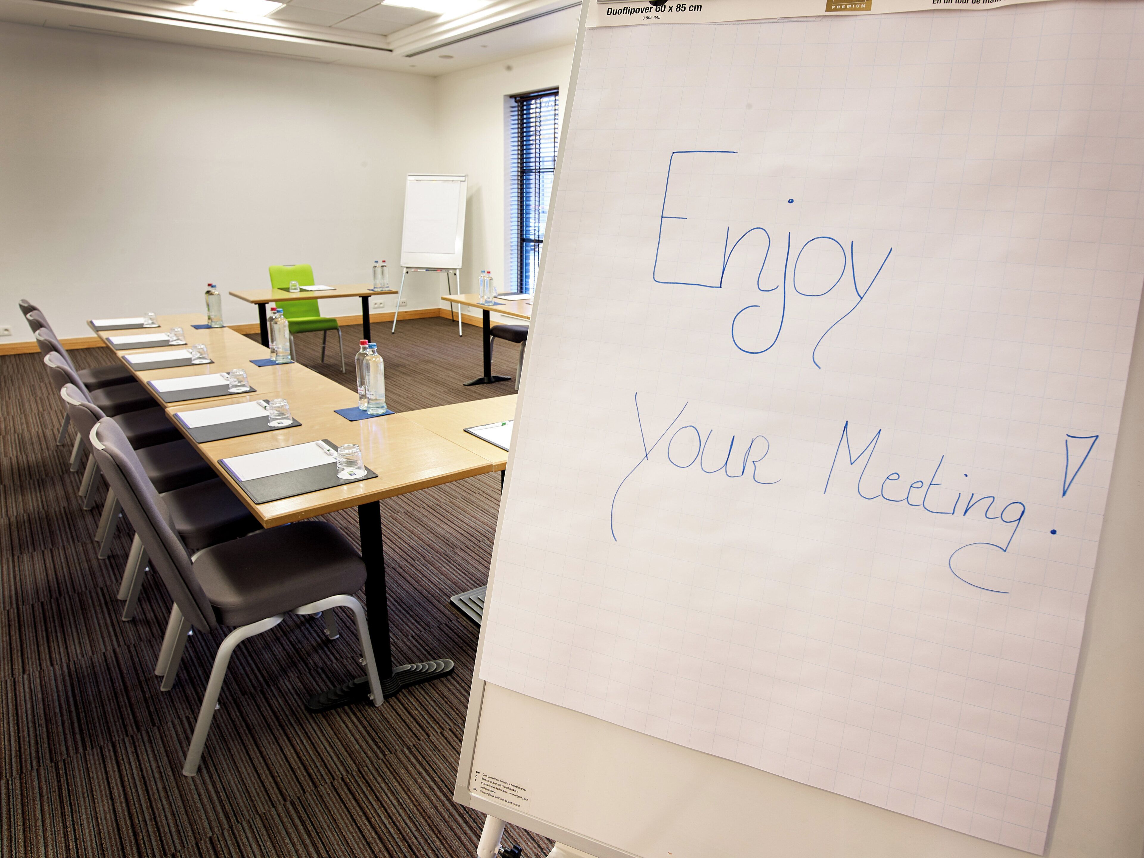 A conference room with a whiteboard displaying 'Enjoy Your Meeting!' and a table with chairs and water bottles.