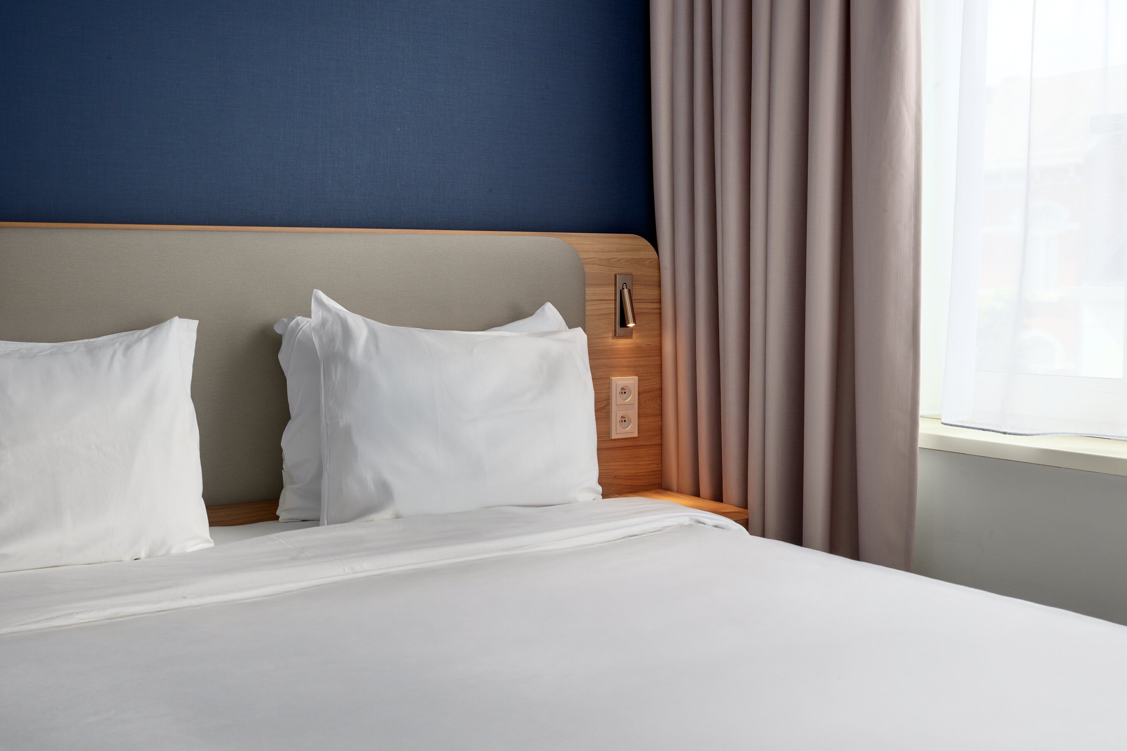 A hotel room with a bed, a wooden headboard, a nightstand, and a window with curtains.