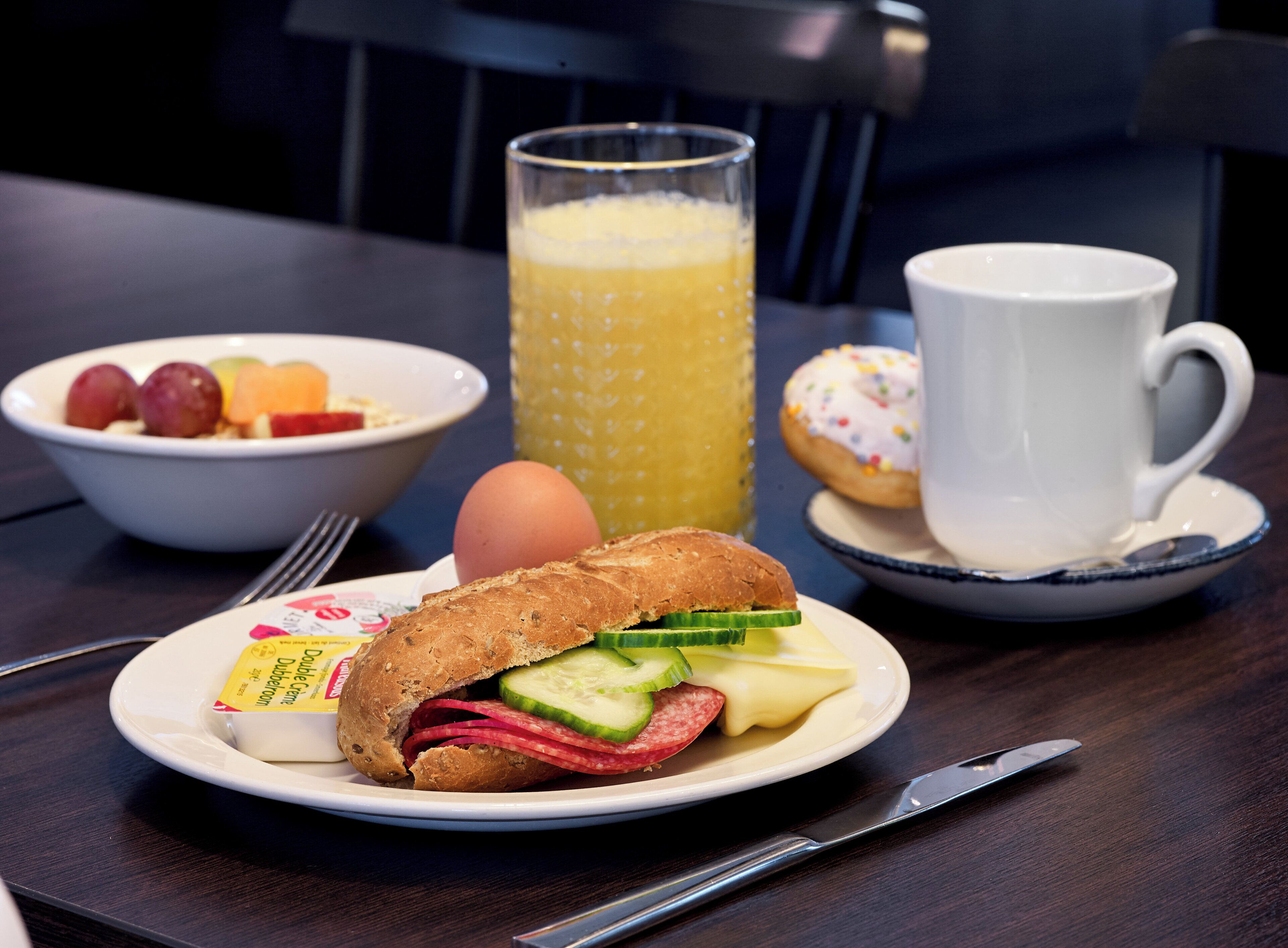 Un desayuno con un sándwich, un bol de fruta, un vaso de jugo de naranja, un muffin y una taza en una mesa.