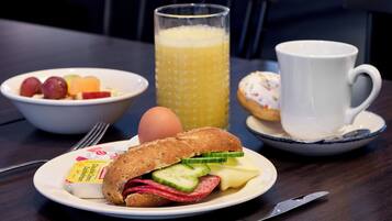 Un desayuno con un sándwich, un bol de fruta, un vaso de jugo de naranja, un muffin y una taza en una mesa.