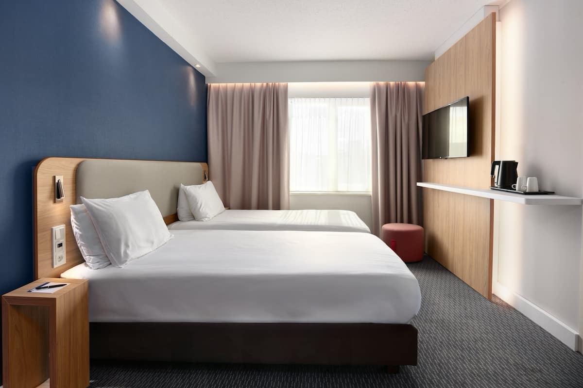 A hotel room with two beds, a TV, a coffee maker, and a small red ottoman.