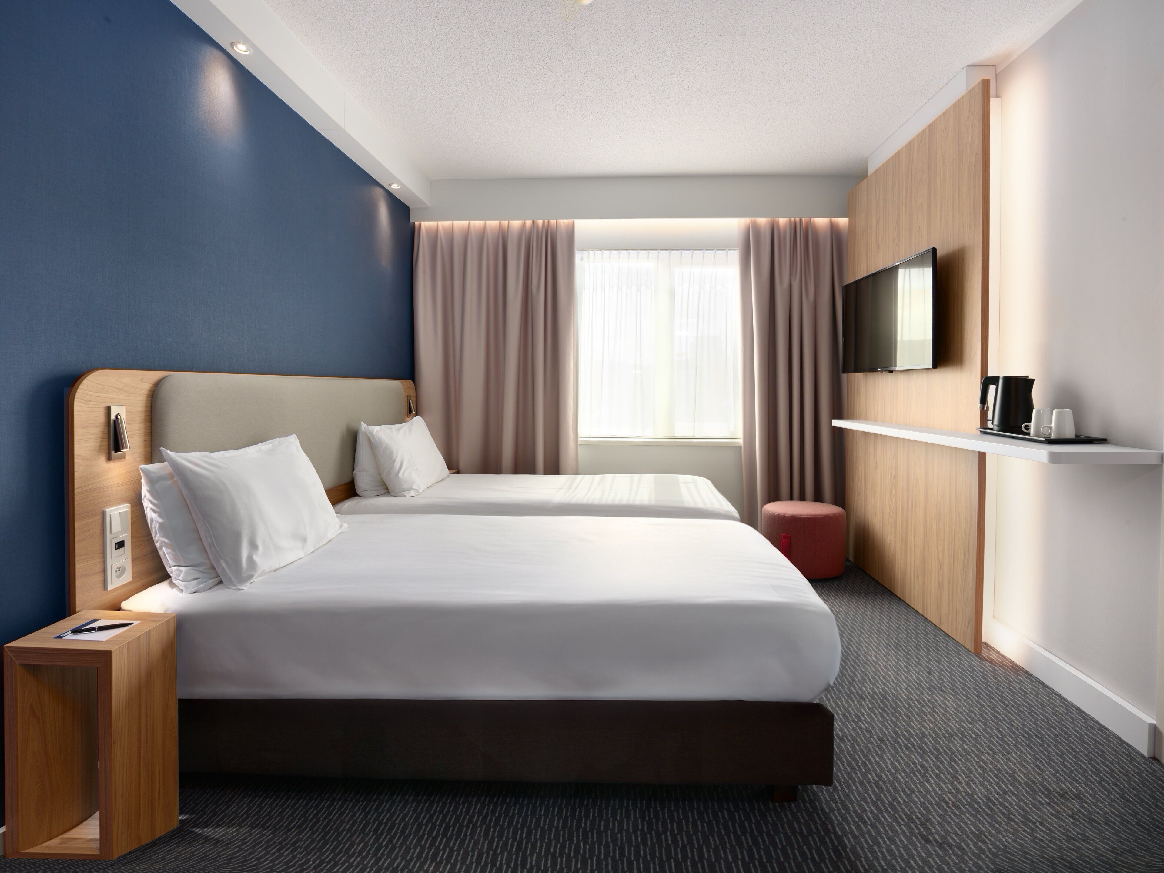 A hotel room with two beds, a TV, a coffee maker, and a small red ottoman.
