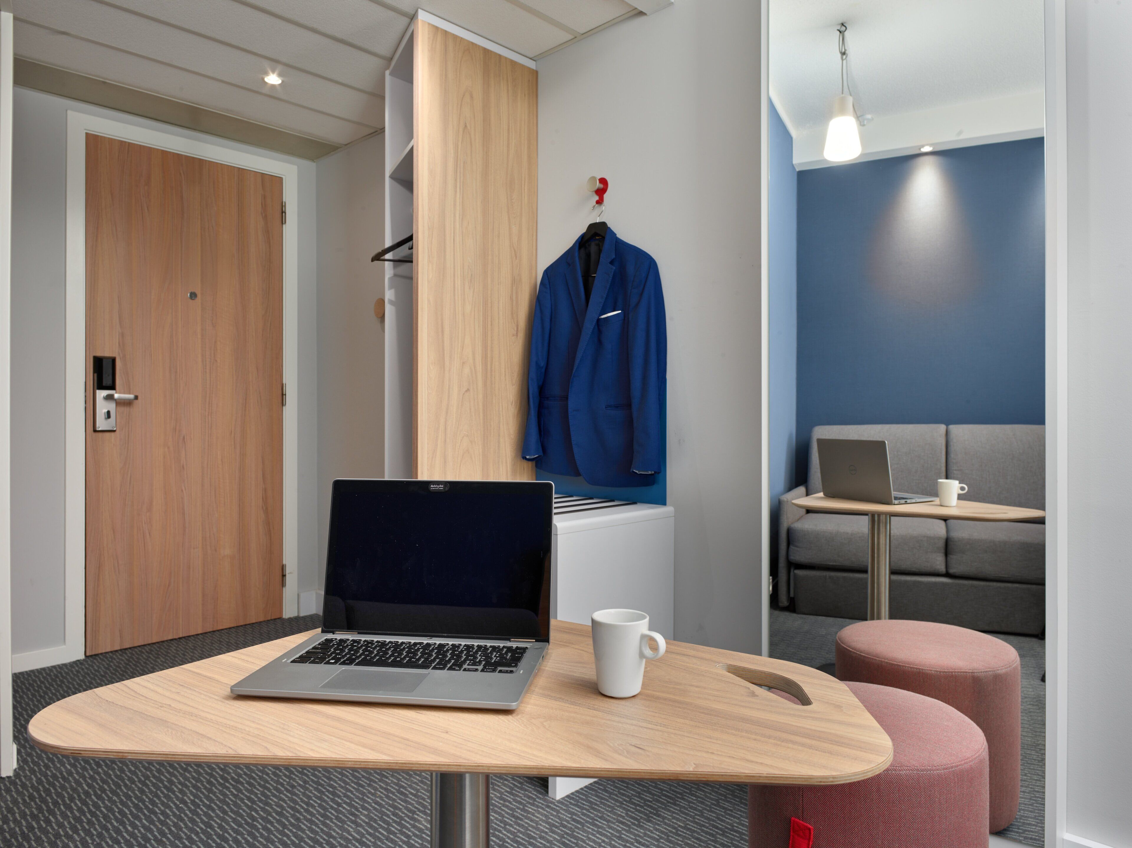 A modern office space with a laptop on a wooden table, a white mug, a blue robe hanging on a rack, and a grey sofa with a laptop and a white mug on a small table.