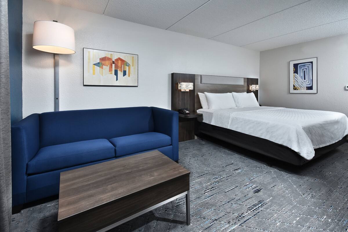 A modern hotel room with a blue sofa, a bed with white bedding, a wooden coffee table, and abstract artwork on the walls.