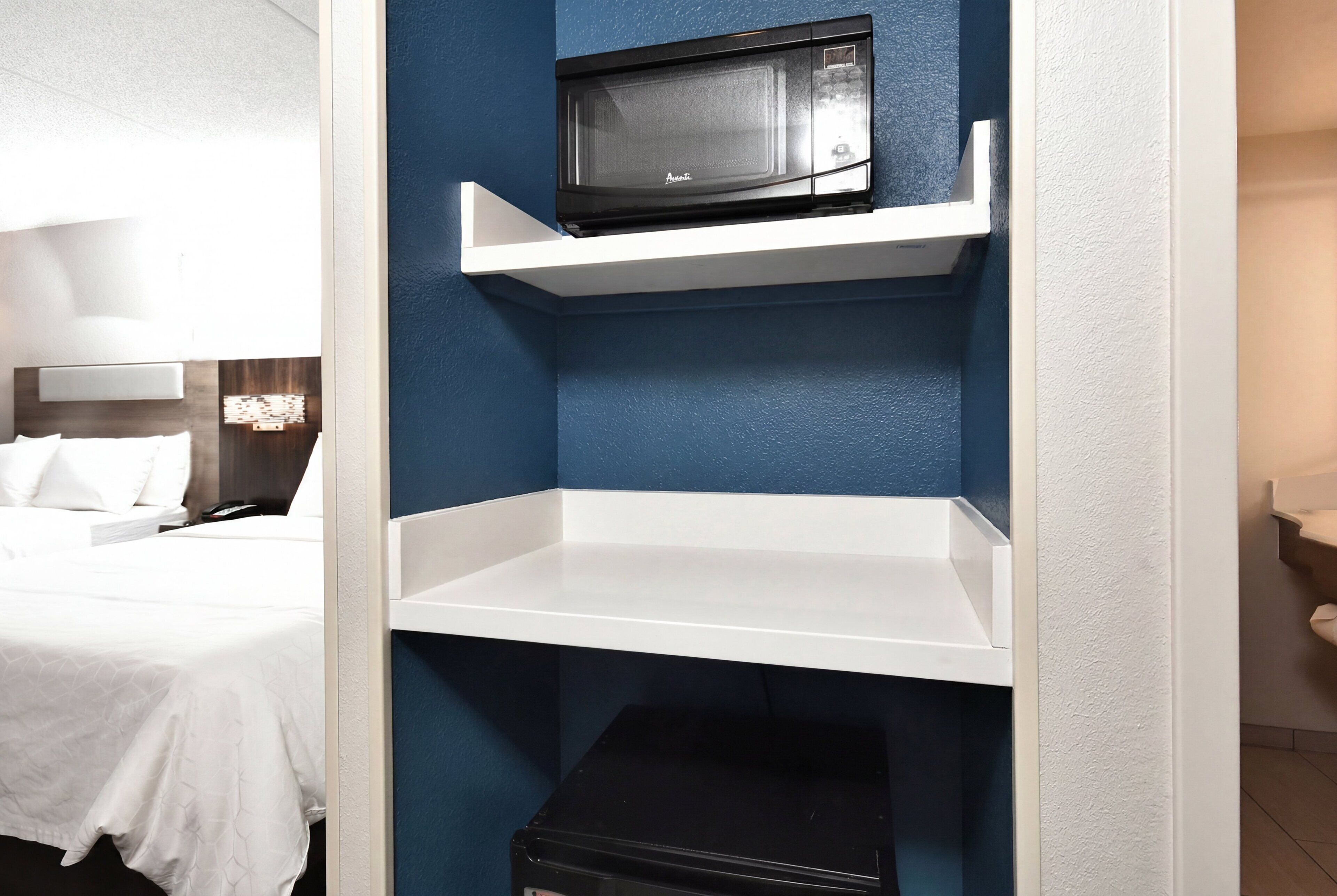 Hotellihuone, jossa on sänky, valkoinen hylly, jonka päällä on mikroaaltouuni, ja musta säilytysyksikkö.