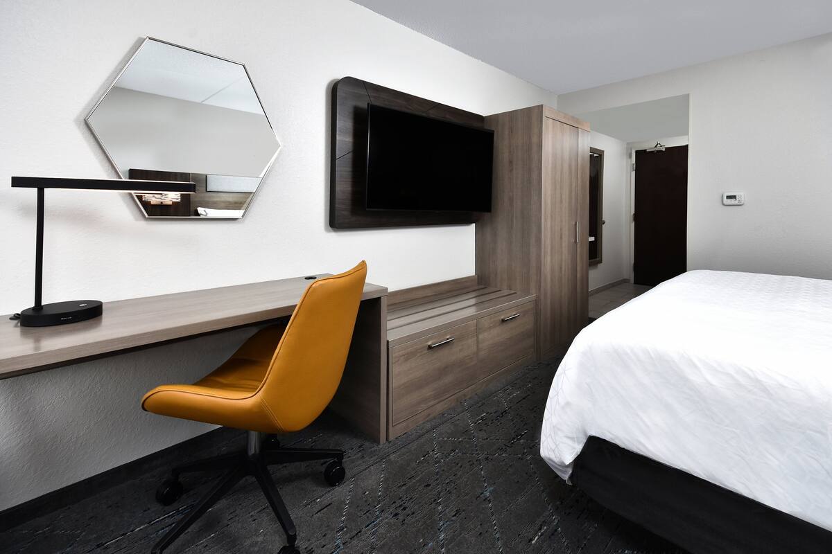 A hotel room with a bed, a desk with a chair, a TV mounted on the wall, and a wardrobe.