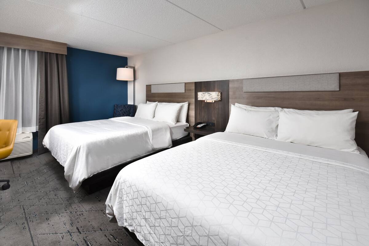 A hotel room with two beds, a desk, a chair, and a lamp.