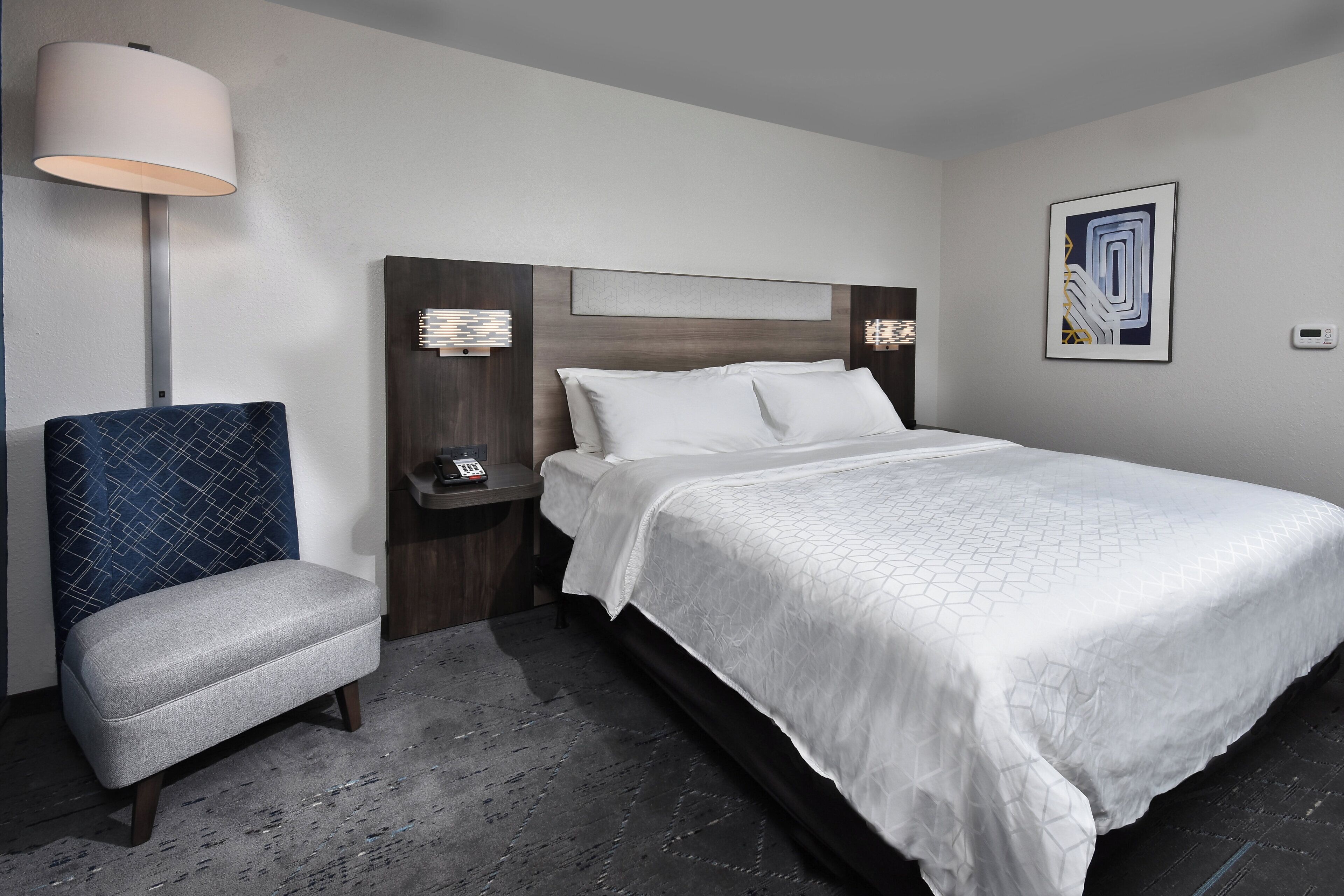 A hotel room with a bed, a chair, a bedside table with a phone, and a wall-mounted light fixture.