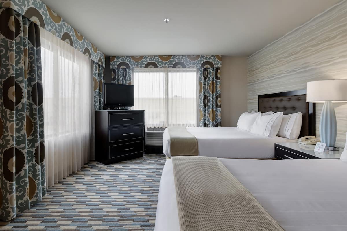A hotel room with a large bed, a bedside table with a lamp, a dresser, a television, and a window with curtains.