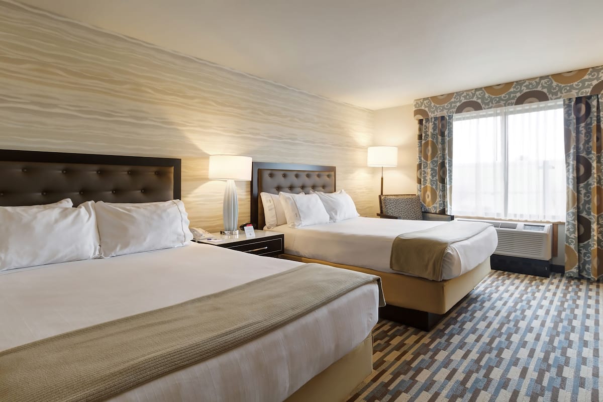 A hotel room with two beds, a large window with curtains, a wall-mounted air conditioner, and a patterned floor.