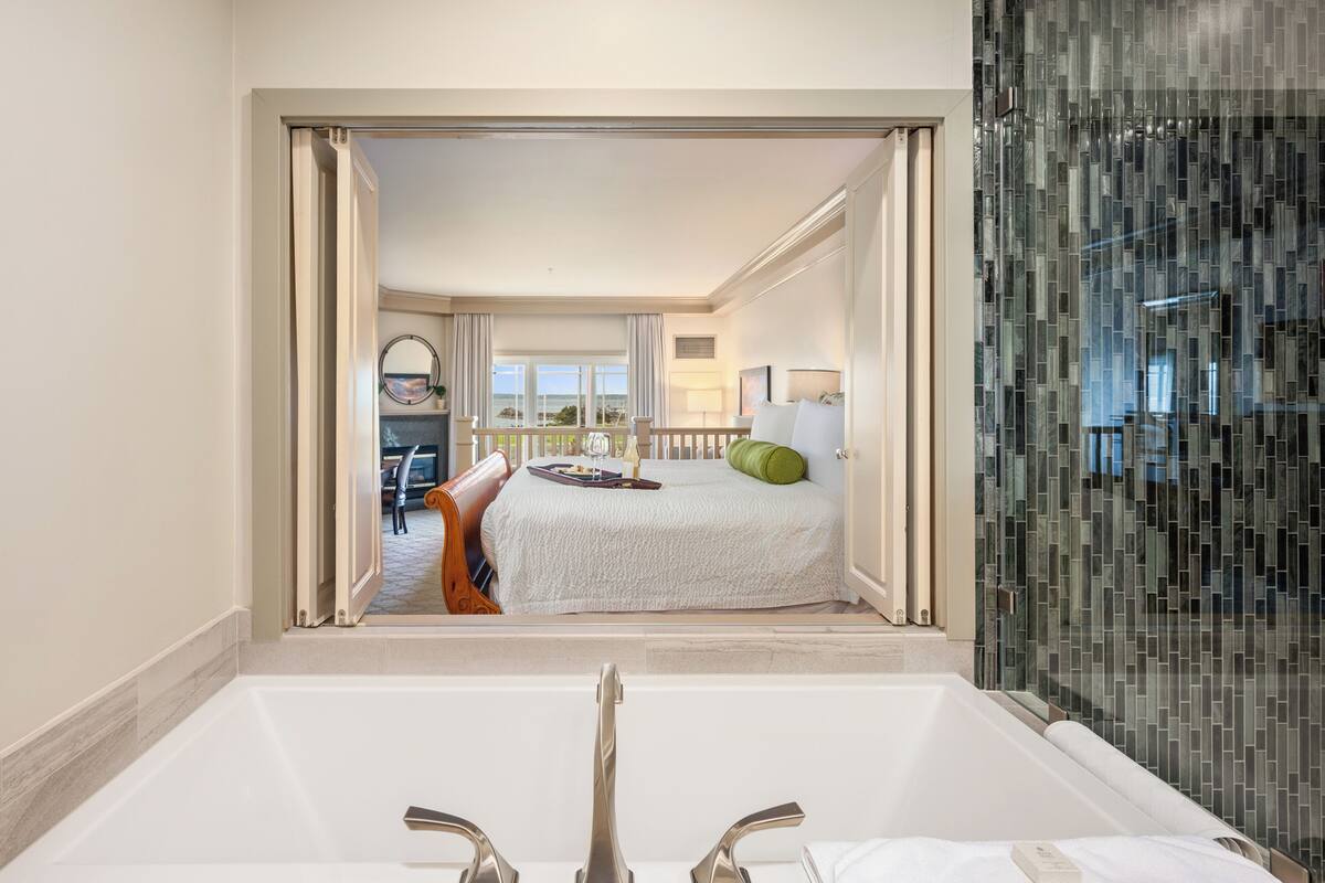 A hotel room with a large bathtub, a bed with a white bedspread, a green pillow, and a view of the outside.