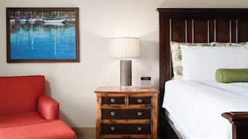 A hotel room with a bed, a nightstand with a lamp, a red armchair, and a painting of a harbor.