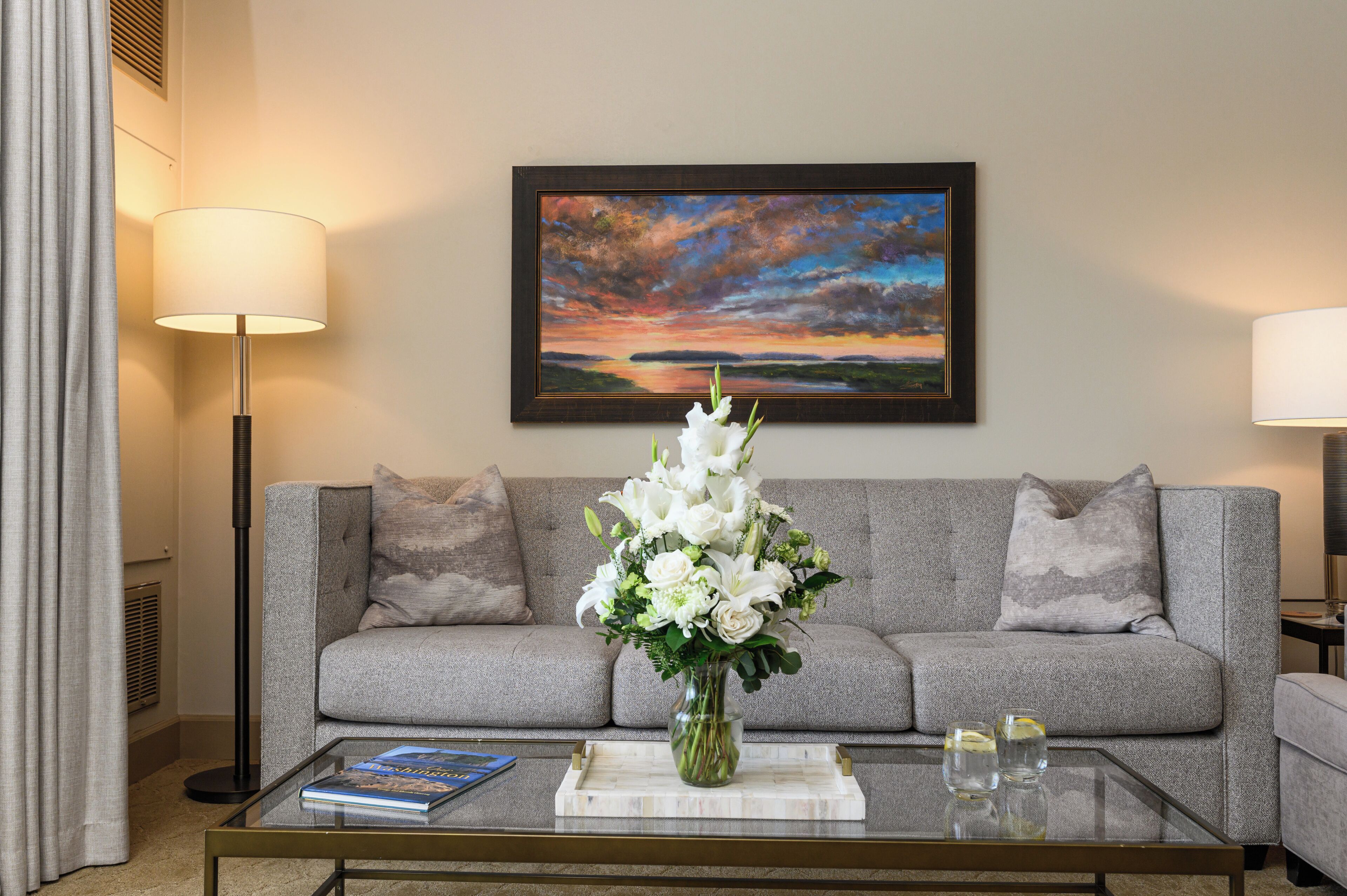 A living room with a grey sofa, a glass coffee table with a book and a vase of flowers, and a painting of a sunset on the wall.