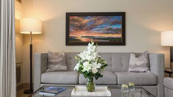 A living room with a grey sofa, a glass coffee table with a book and a vase of flowers, and a painting of a sunset on the wall.
