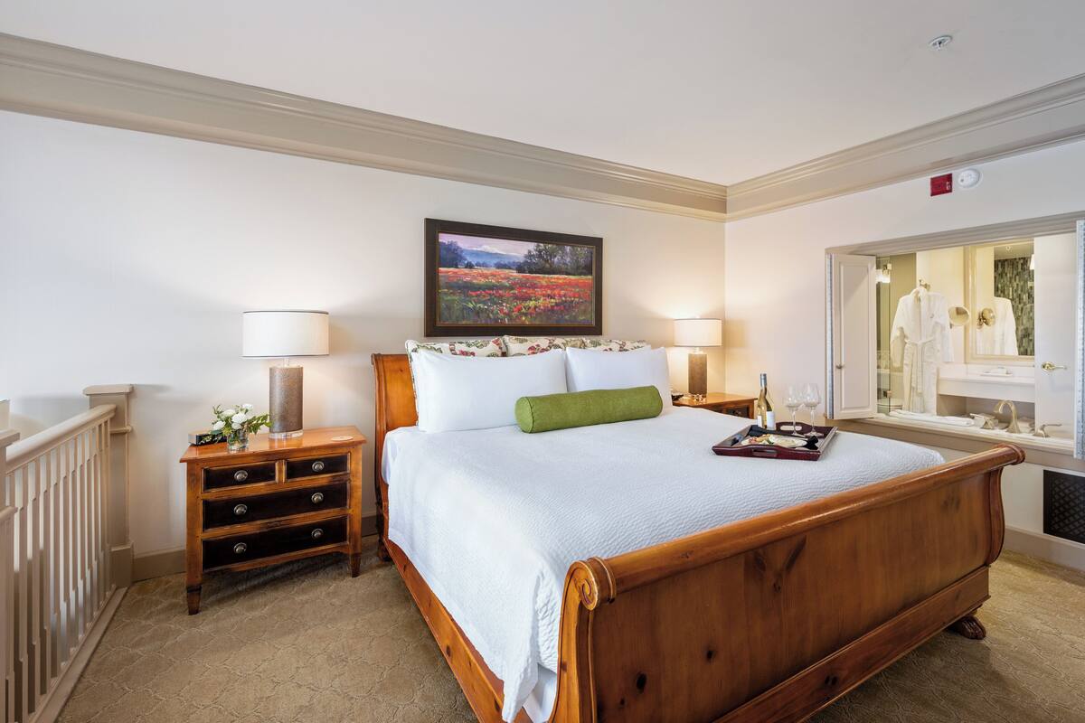 A neatly arranged bedroom with a wooden bed, a nightstand with a lamp, a framed painting, and a bathroom visible through an open door.
