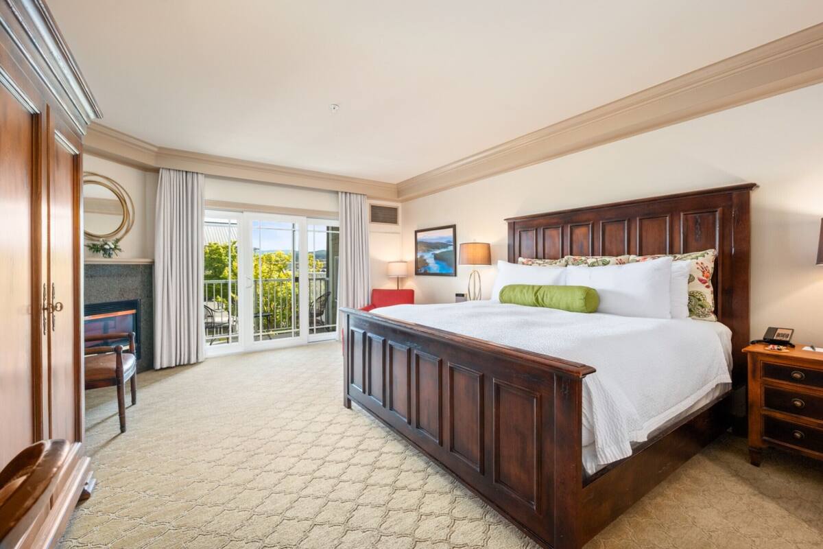 A bedroom with a large bed, a wooden nightstand, a desk with a chair, a large window with curtains, and a view of greenery outside.